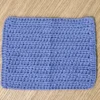 A single periwinkle blue crochet washcloth shown flat on a wooden surface, highlighting the uniform textured stitch and rectangular shape.
