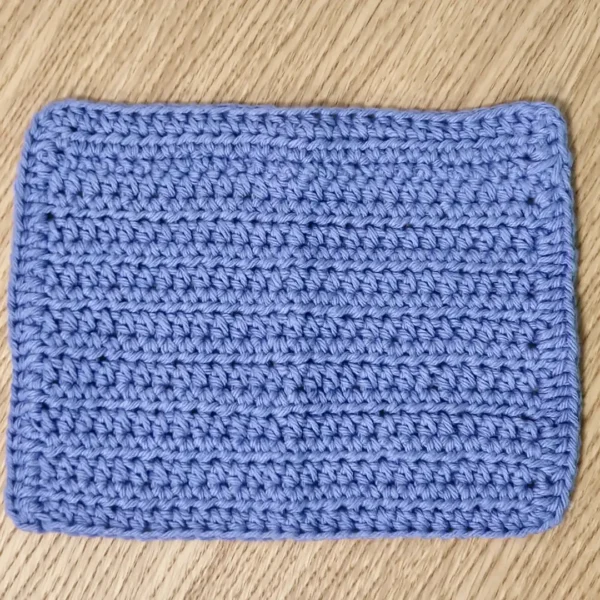 A single periwinkle blue crochet washcloth shown flat on a wooden surface, highlighting the uniform textured stitch and rectangular shape.