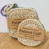 A periwinkle-lilac round crochet face disc resting against a sand-colored square scrubby, both featuring Patient Calf branding.