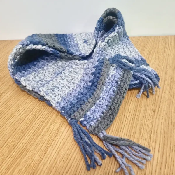The Drina Scarf by Patient Calf, showing blue and grey crochet stripes and tassel ends on a light surface.
