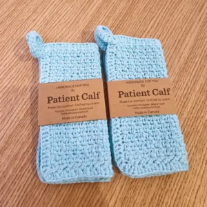 Two hand-crocheted light blue cotton washcloths with hanging loops by Patient Calf, featuring a textured stitch for skincare exfoliation.